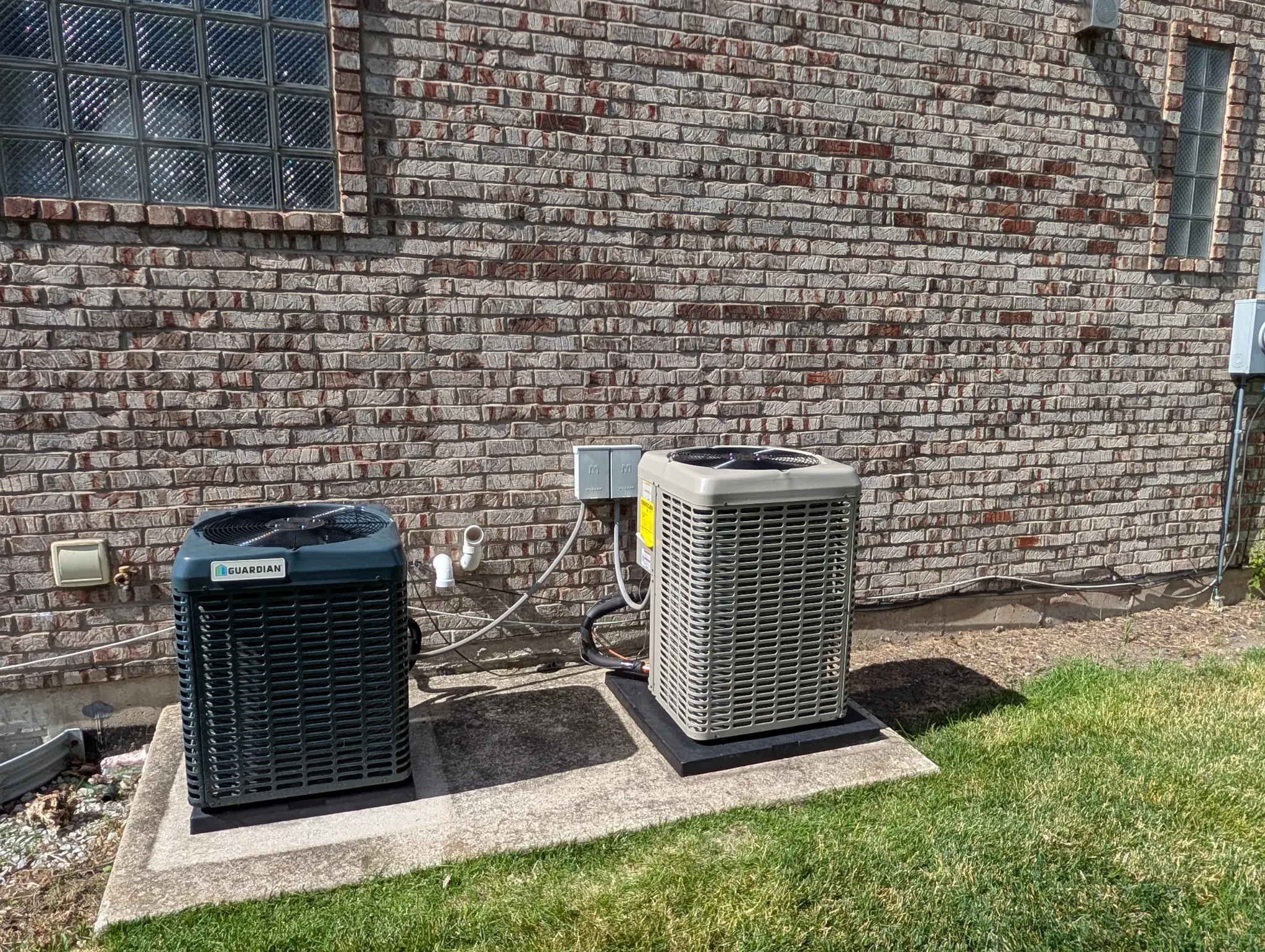 Two outdoor HVAC condensers on pads beside a brick wall